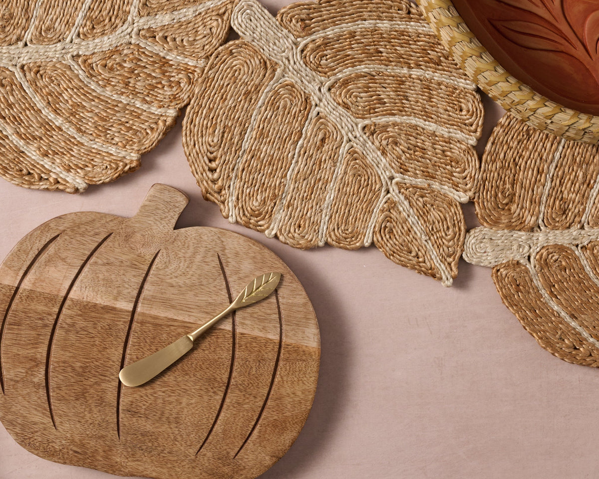 A styled flat-lay of woven home decor and serving pieces on a dusty pink background, featuring a leaf-shaped woven runner, a carved wooden pumpkin serving board with a small gold-toned knife, and a bread warming basket with terracotta insert.