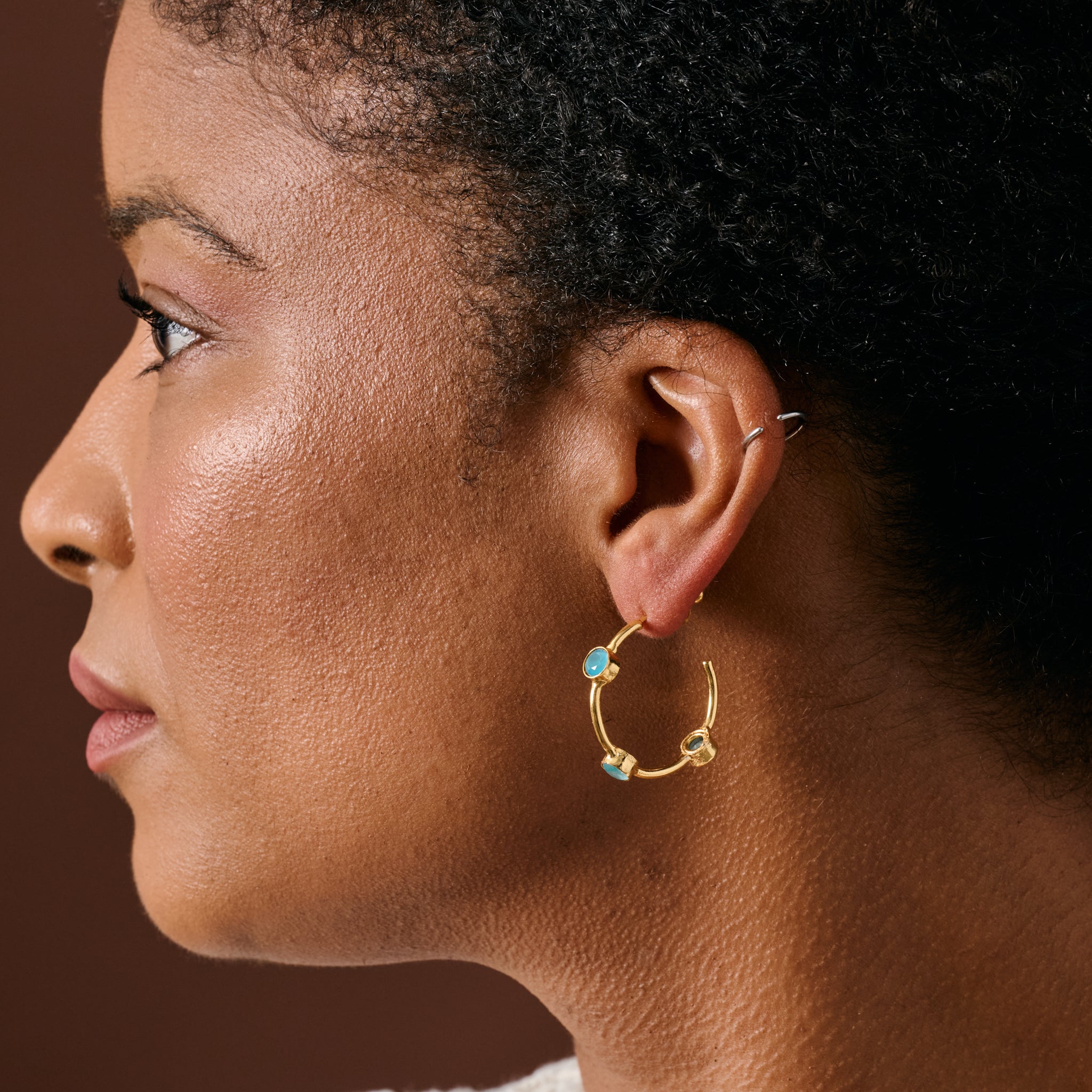 A close-up side profile of a woman wearing a delicate gold hoop earring adorned with small light blue gemstones, set against a dark brown background.