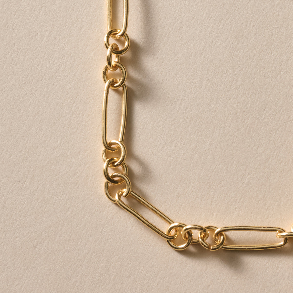 A close-up of a gold-toned chain necklace with elongated oval links and smaller connecting rings, laid on a warm off-white textured surface.