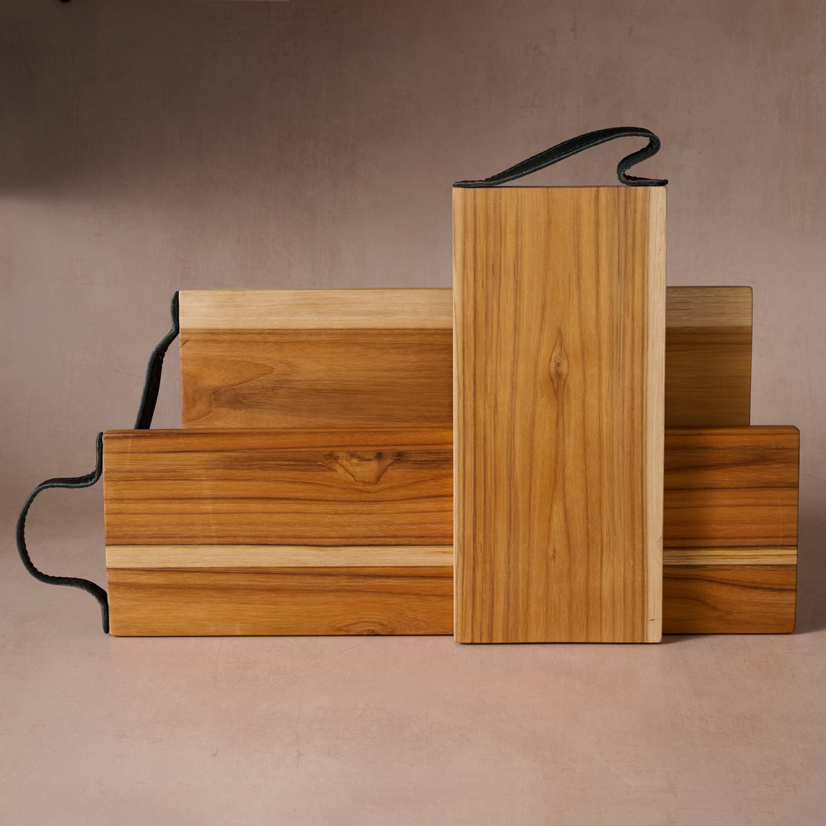 Three handcrafted teak wood cutting boards in assorted sizes with natural wood grain and leather hanging handles.