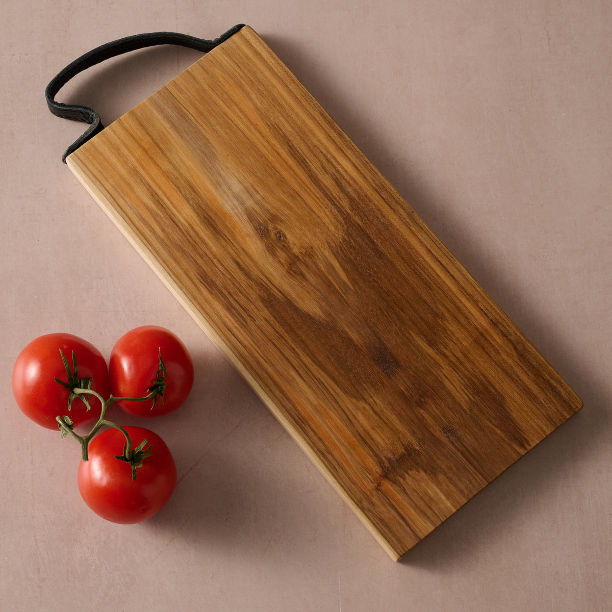 Handcrafted teak wood cutting board with natural wood grain and leather hanging handle, displayed with fresh tomatoes.