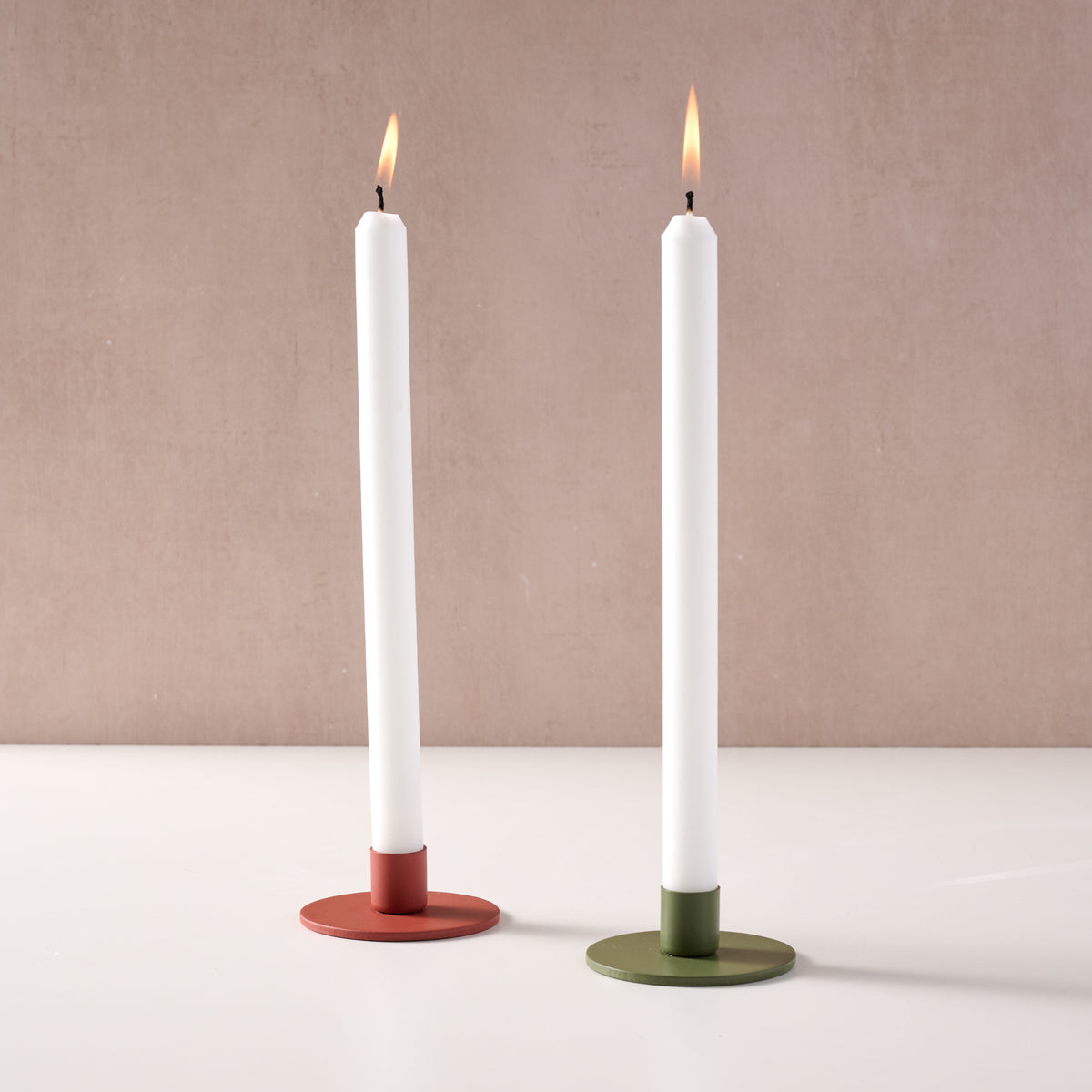 Two tall white taper candles in round metal holders, one with a red base and one with an olive green base, standing on a white surface against a warm beige wall.