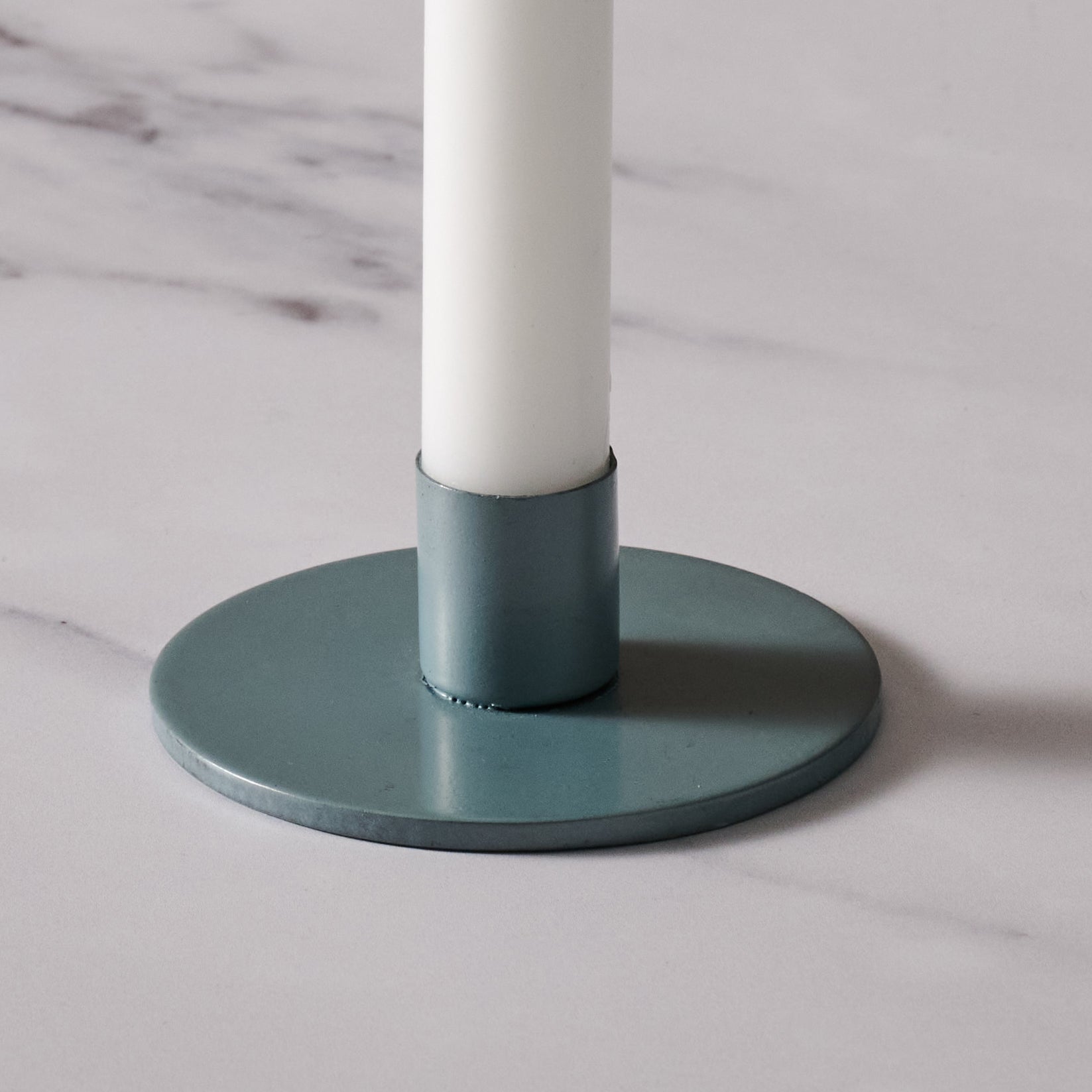 A close-up of a round blue/gray metal candle holder base with a matching teal cylindrical holder, holding a white candle on a light marble surface.