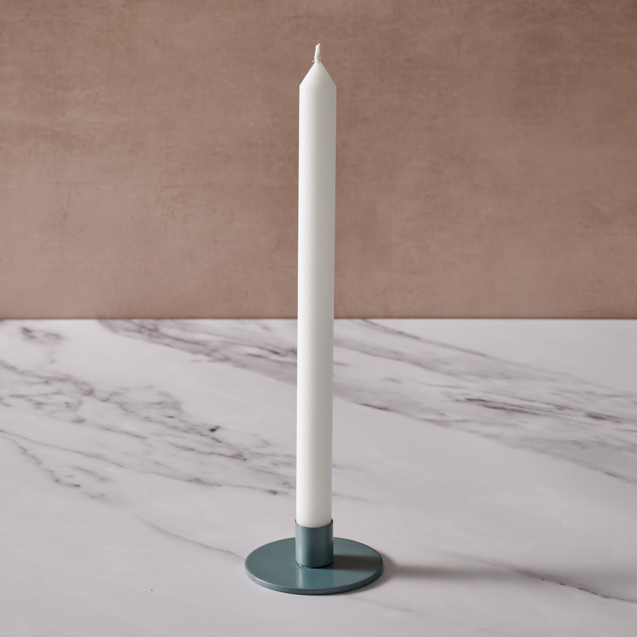 A tall white taper candle in a round blue/gray metal holder, set on a white marble surface against a textured beige-brown background.