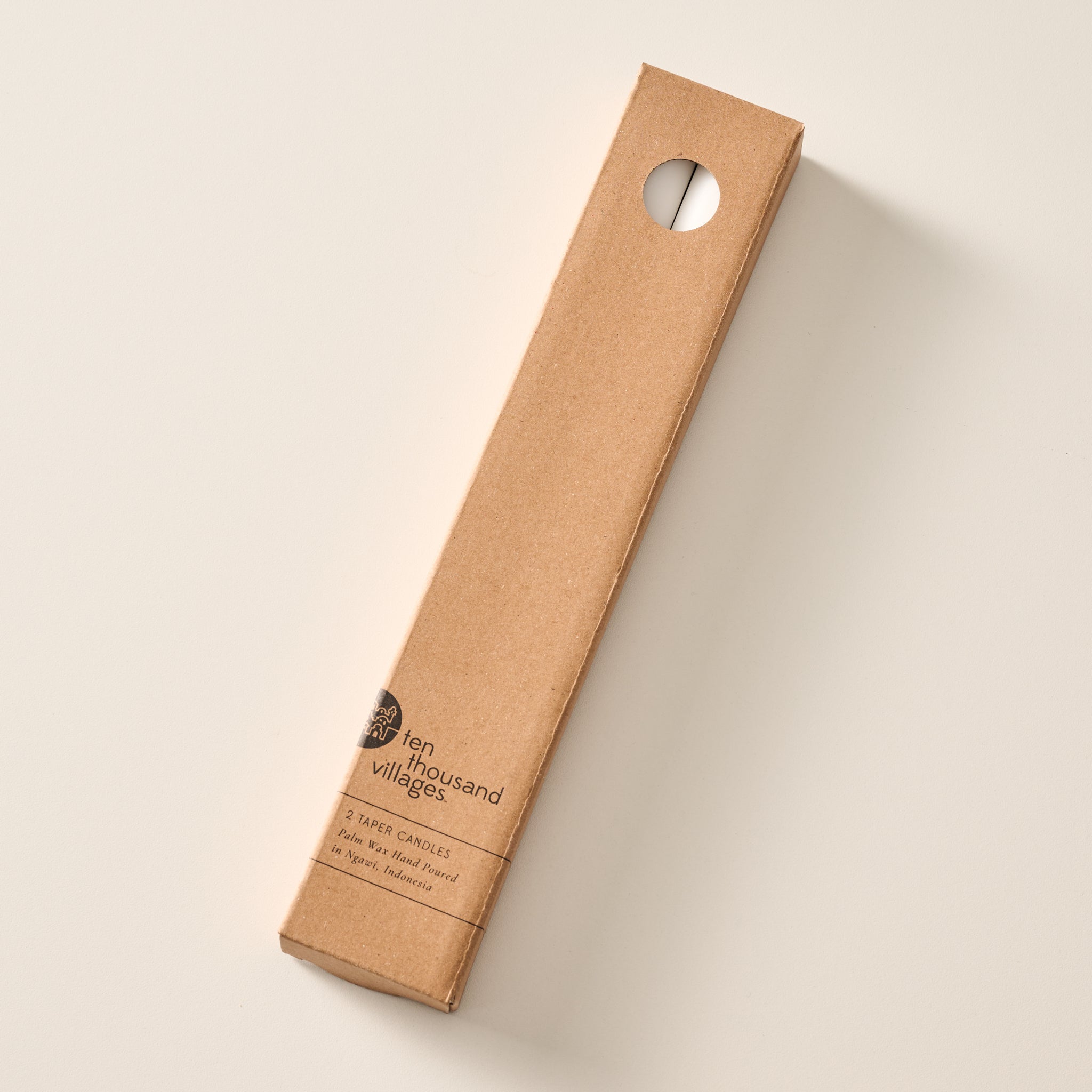 A tall, narrow cardboard box with a circular window display cutout at the top showing two natural white candles inside, photographed on a clean off-white background. The box has printed text near the bottom including the brand name 'Ten Thousand Villages'.
