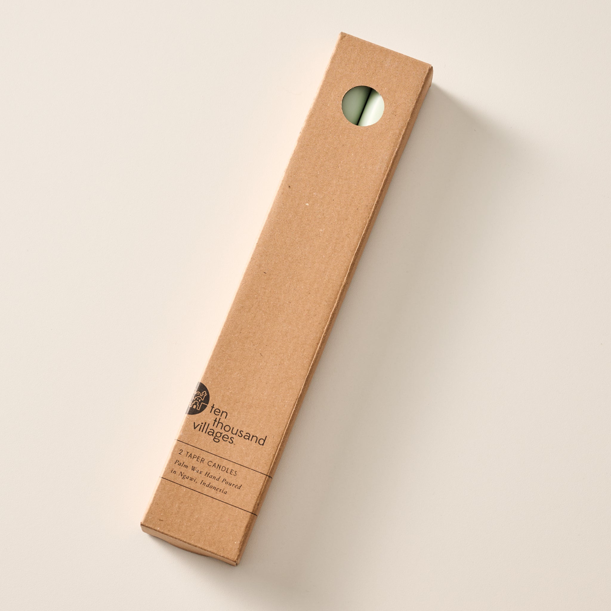 A tall, narrow cardboard box with a circular window display cutout at the top showing two sage green candles inside, photographed on a clean off-white background. The box has printed text near the bottom including the brand name 'Ten Thousand Villages'.