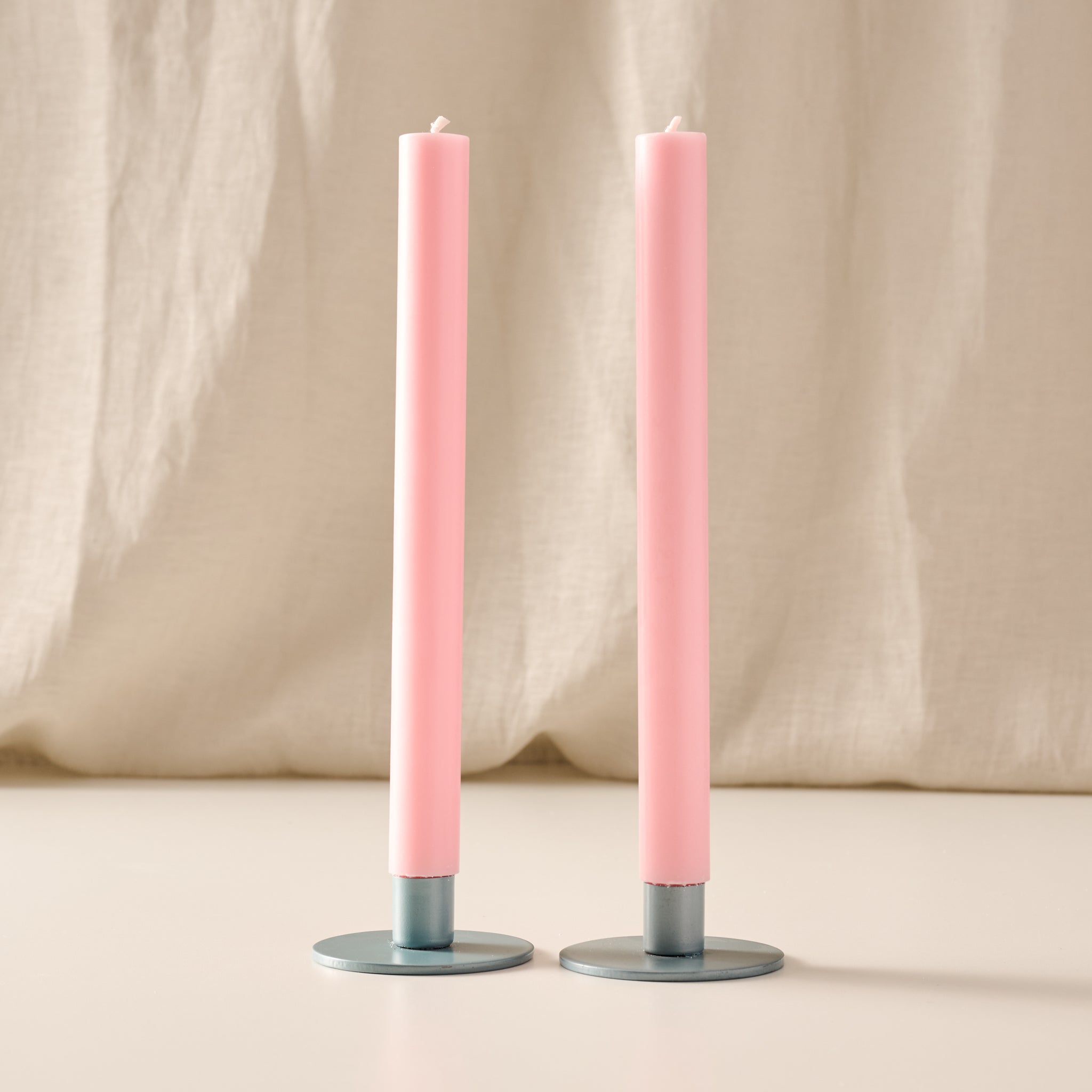 Two tall ginger pink colored taper candles standing upright in round blue/gray metal holders against a soft beige fabric backdrop.
