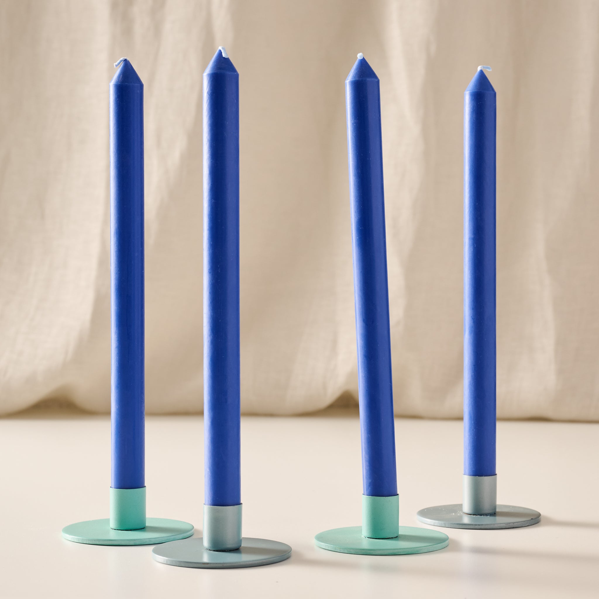 Four cobalt blue taper candles with turquoise and blue/gray candle holders, arranged on a light surface against a soft beige fabric backdrop.