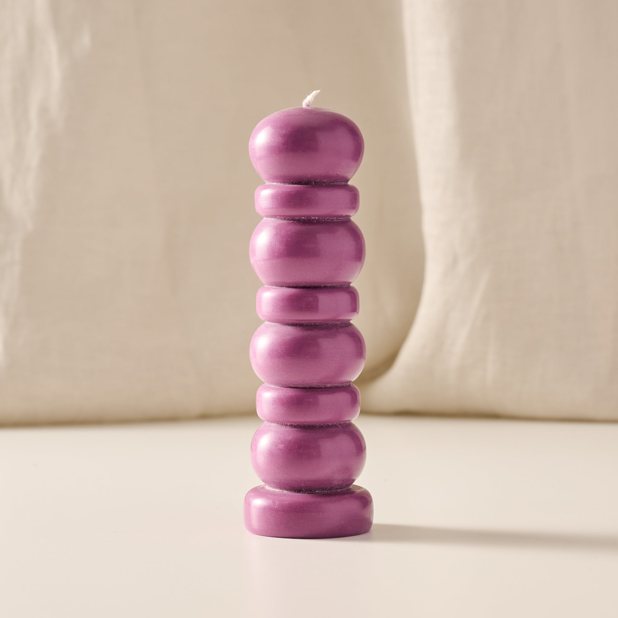 A single purple stacked bead-style candle with a small white wick, standing upright on a light beige surface against a soft beige fabric backdrop.