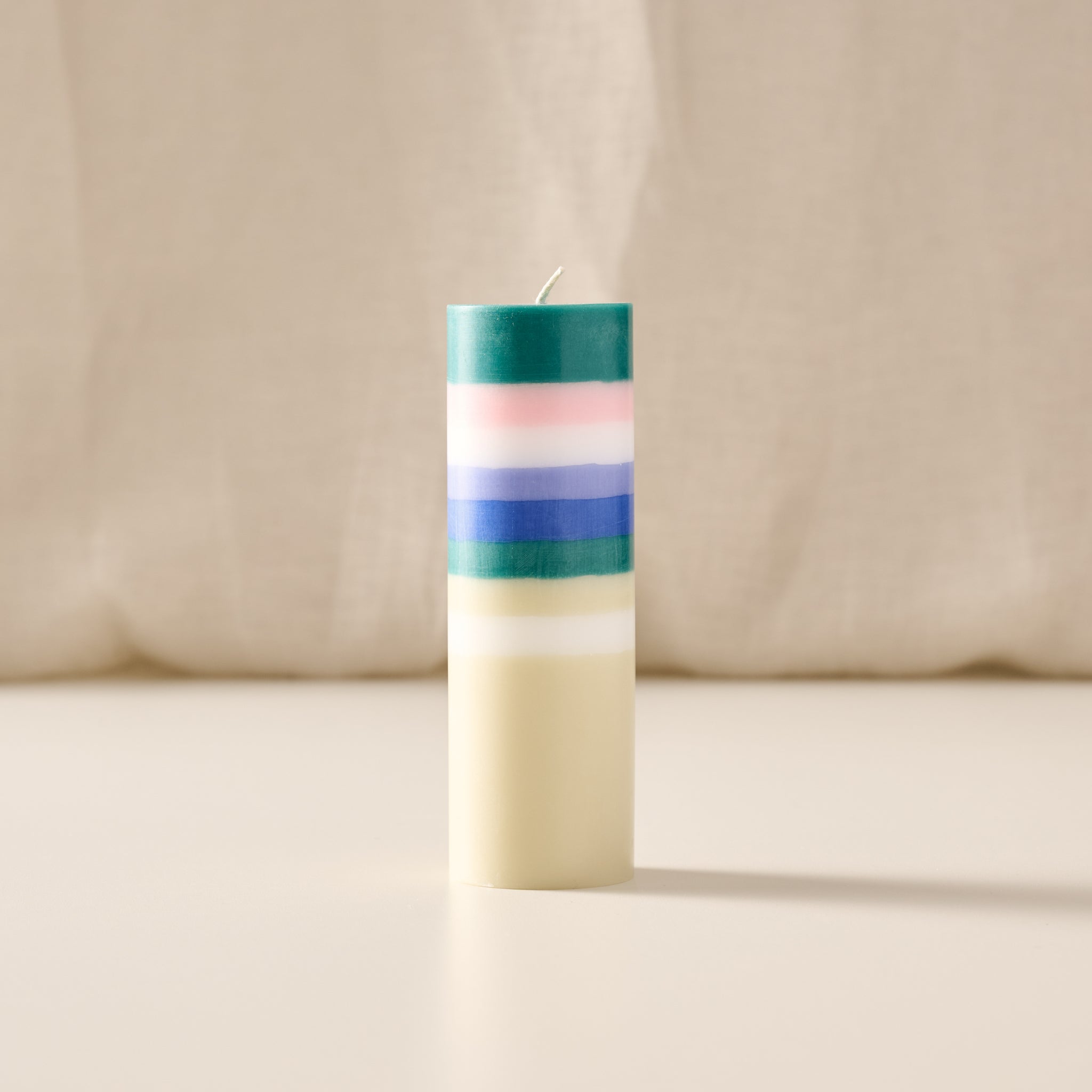 A tall, cylindrical candle with horizontal stripes in teal, pink, white, and blue, with the lower half in solid cream-colored wax, set against a soft beige fabric backdrop.