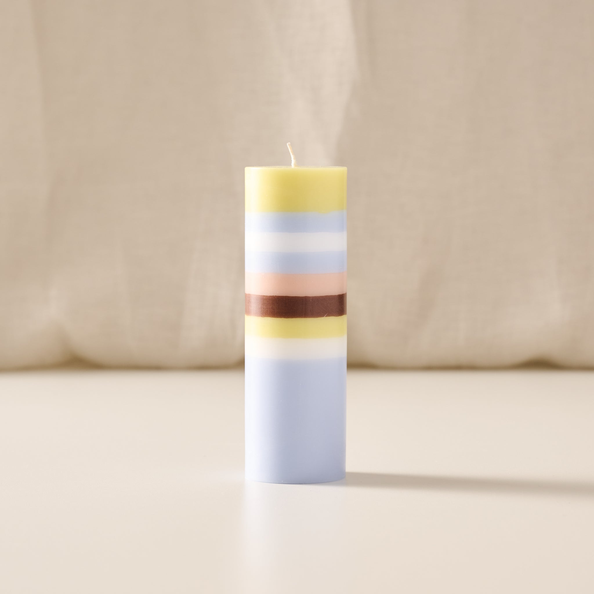 A tall, cylindrical striped candle with pastel yellow, light blue, white, peach, and dark brown bands, standing upright on a light beige surface against a soft beige fabric backdrop.