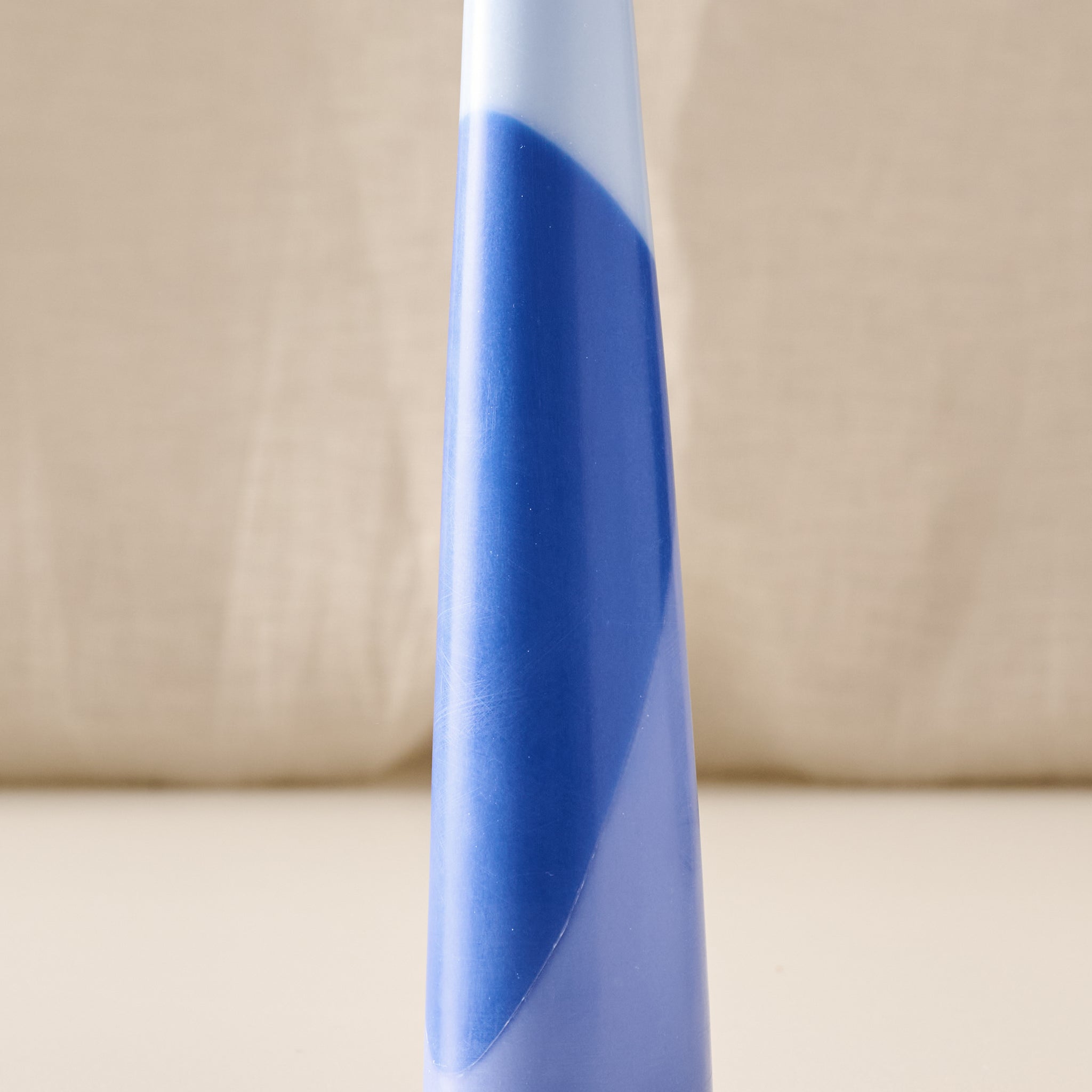 A close-up of a tapered candle with a blue and light blue geometric pattern, set against a neutral beige fabric background.