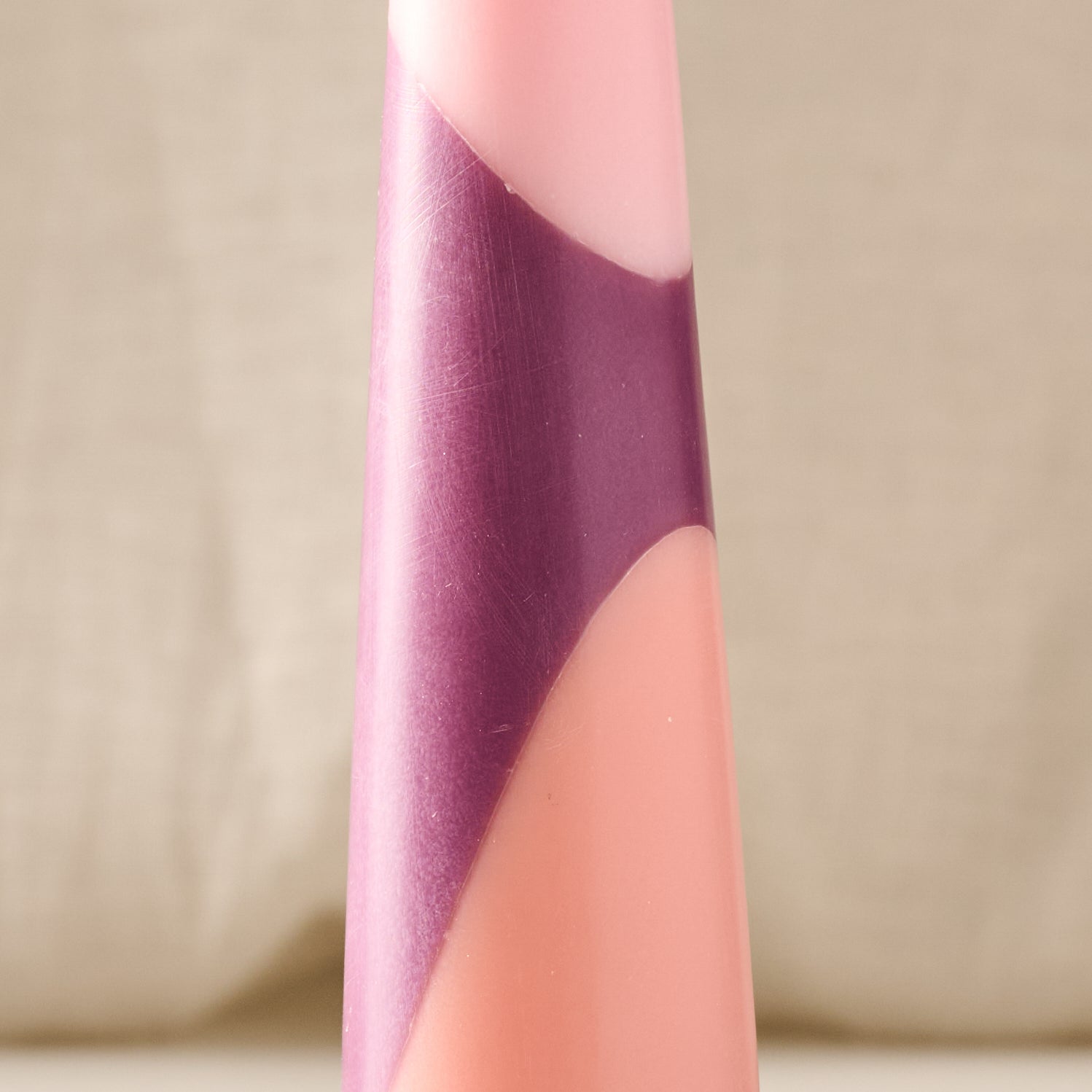 A close-up of a tapered candle with a diagonal color-blocked design in matte dusty pink and metallic purple, set against a soft beige fabric background.