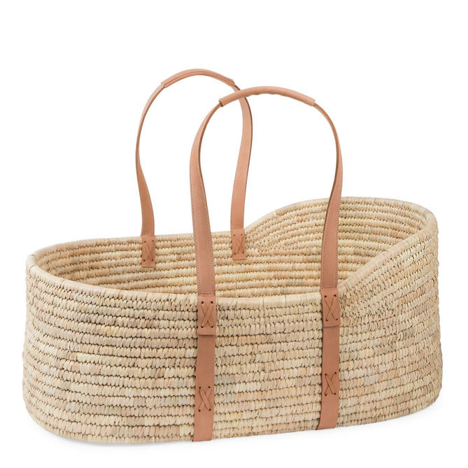 A hand woven moses basket sits in a wooden stand in the corner of a bright room with wood floors, white walls and a tall hand woven hamper next to it. 